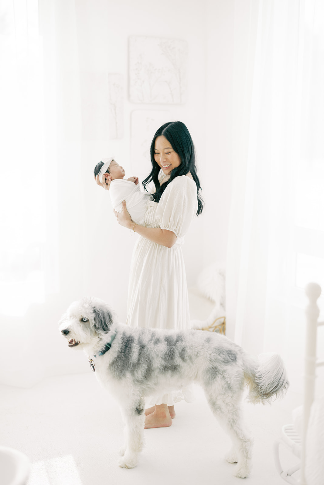 A happy mom stands in a nursery holding her newborn and laughing at her white and grey dog at her side after visiting Minneapolis baby stores