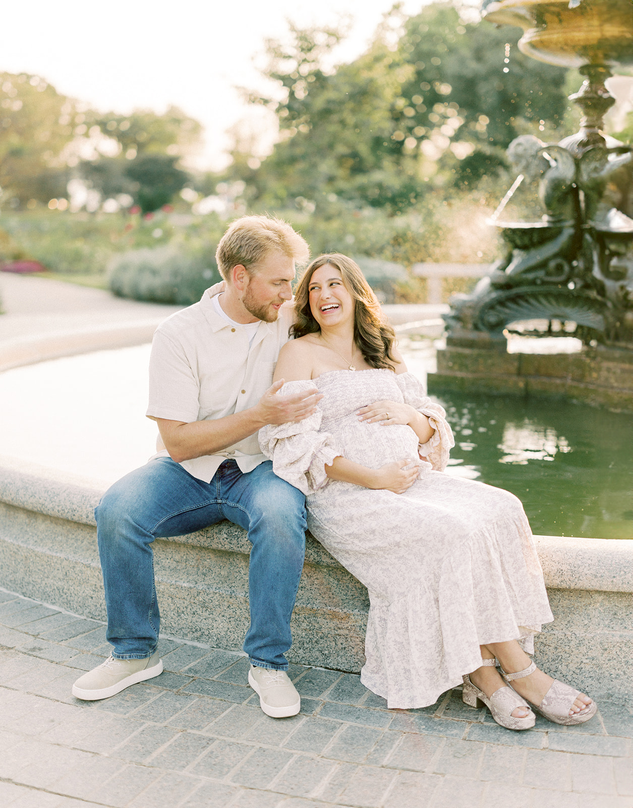 A happy expecting couple sit on a fountain wall snuggling while holding the bump