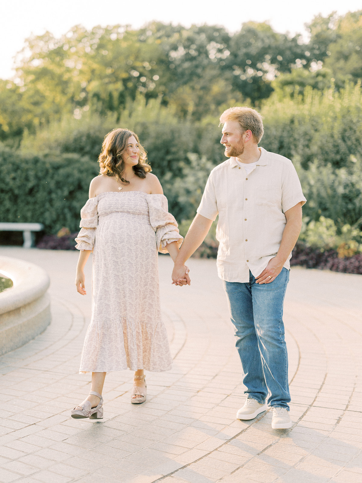 A happy expecting couple strolls in a garden holding hands at sunset after visiting baby shower venues in Minneapolis