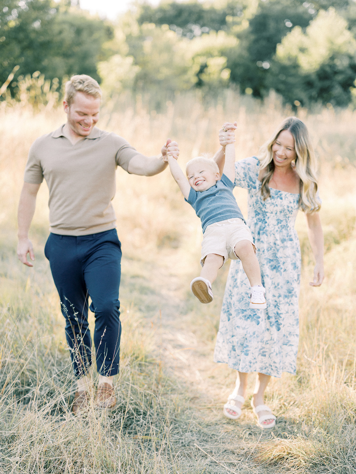 A happy toddler boy swings in mom and dad's hands in a trail of tall grass at sunset