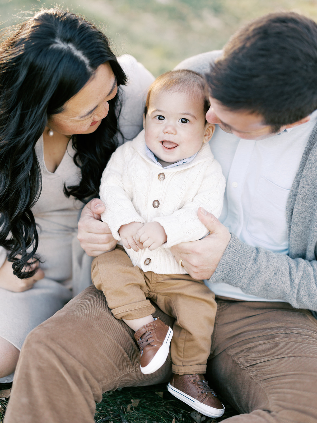 A happy baby in a white sweater sits in mom and dad's lap before visiting indoor playgrounds in Minneapolis