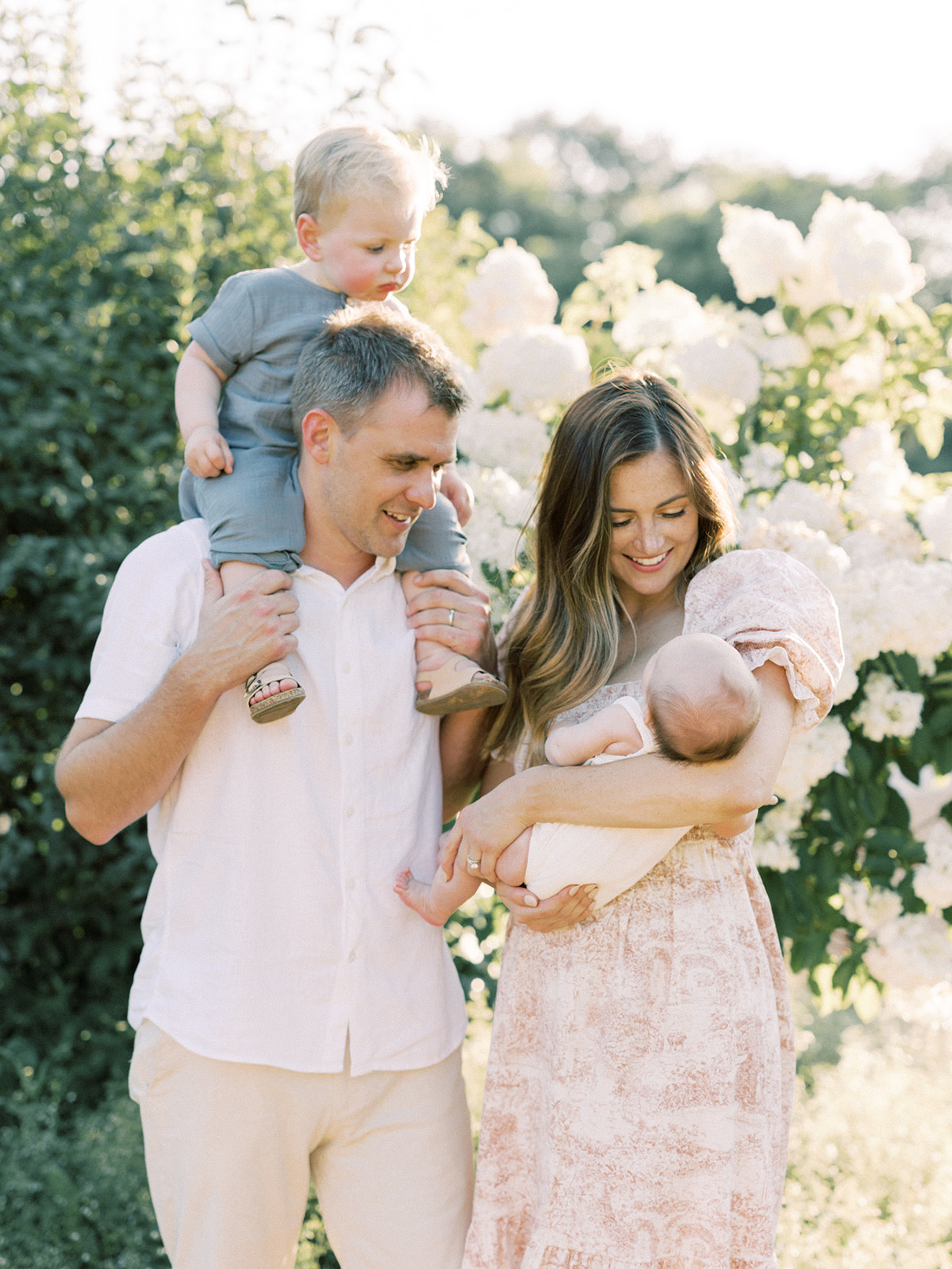 A happy family explores a flower garden at sunset with toddler on dad's shoulders and newborn in mom's arms