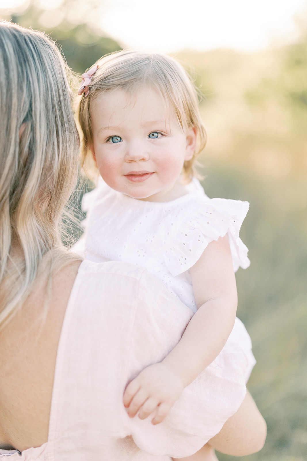 A happy baby in a white dress sits in mom's arms in a park at sunset after using lactation consultants in Minneapolis