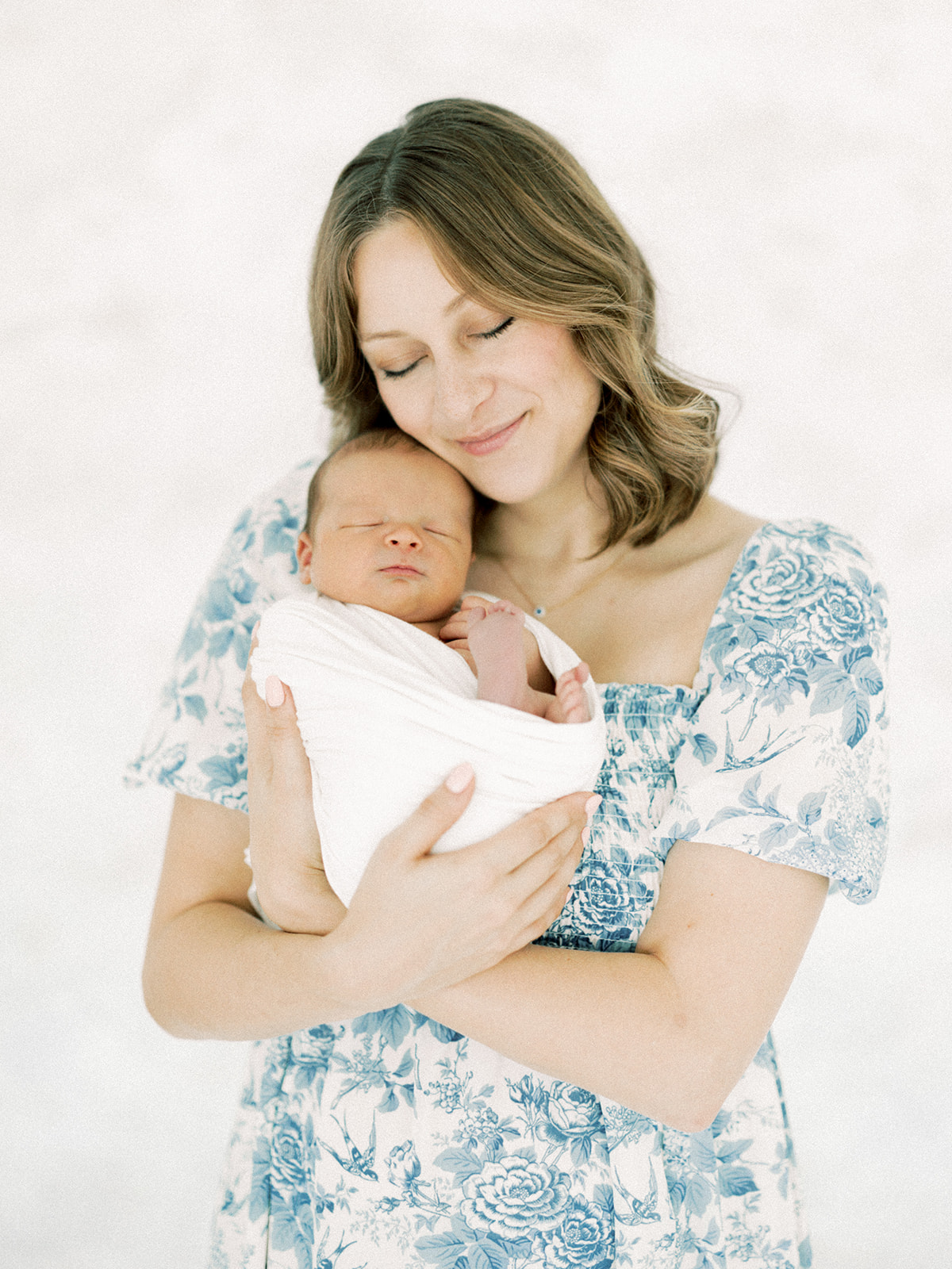 A smiling new mom in a blue floral dress snuggles her sleeping newborn against her cheek