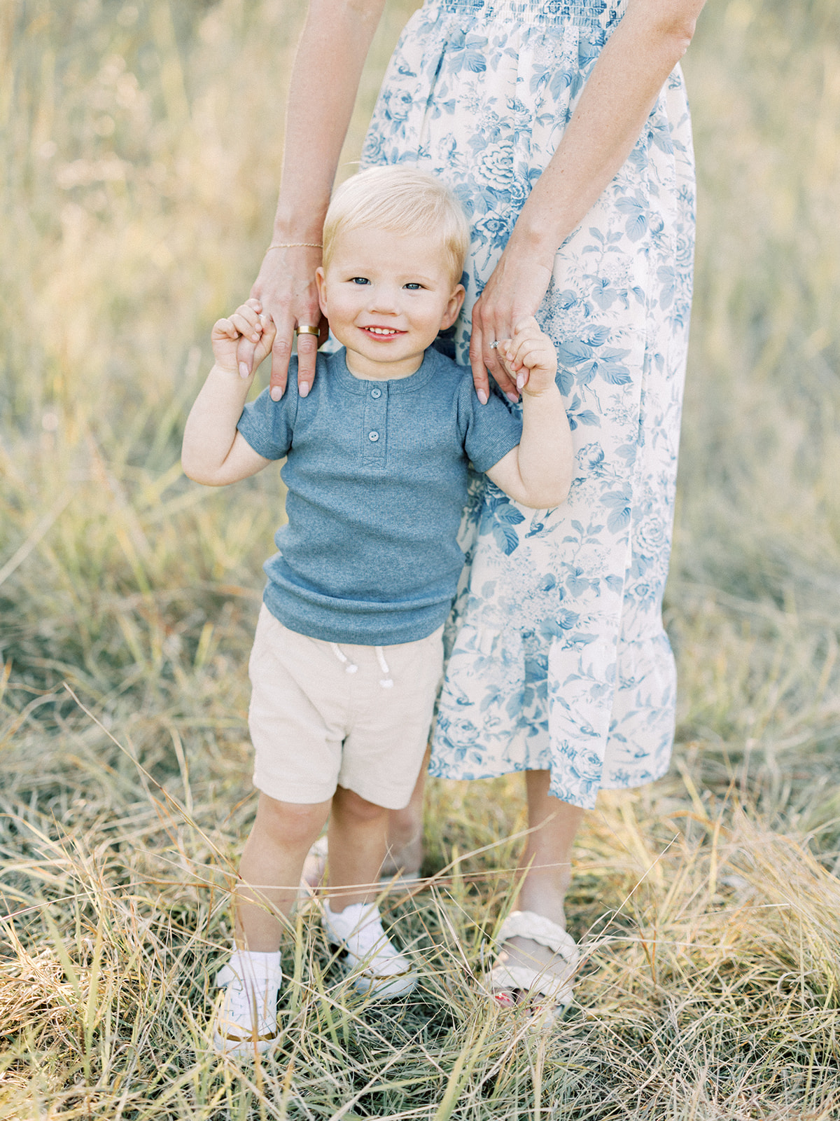 A toddler boy in a blue shirt stands holding mom's hands in a field of tall grass after meeting Minneapolis Midwives