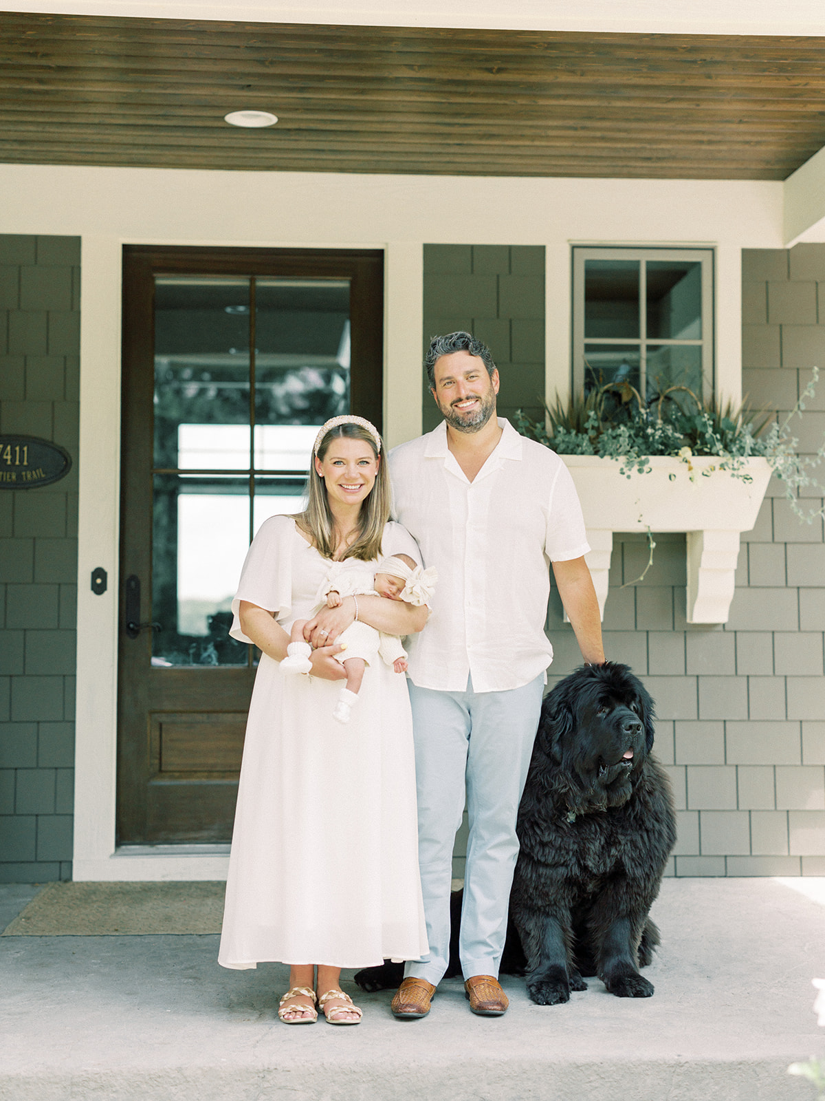 Happy new parents stand on their porch holding a sleeping newborn with their large black dog