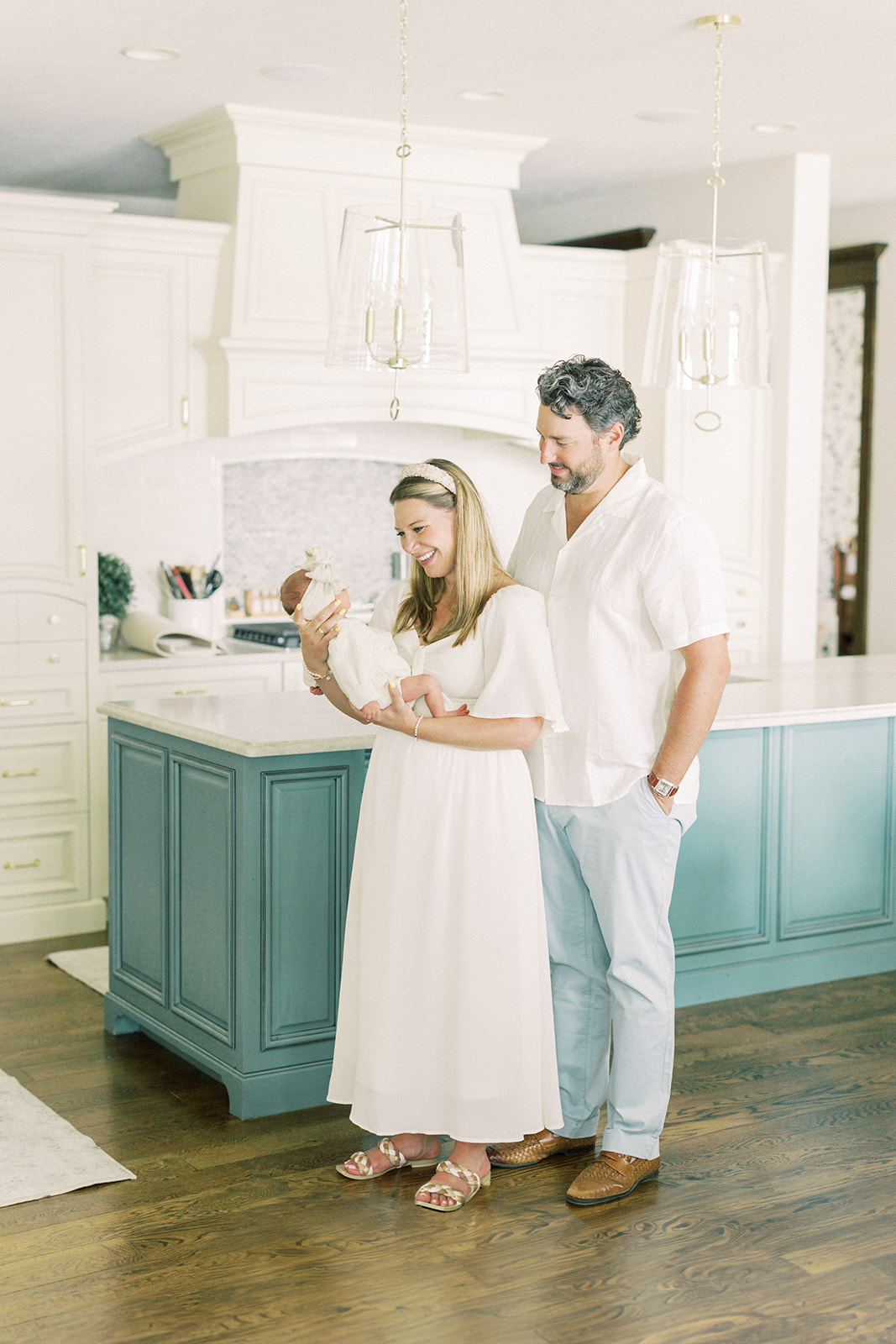 A happy mom and dad stand in a kitchen admiring their sleeping newborn in mom's hands after finding new mom groups in Minneapolis