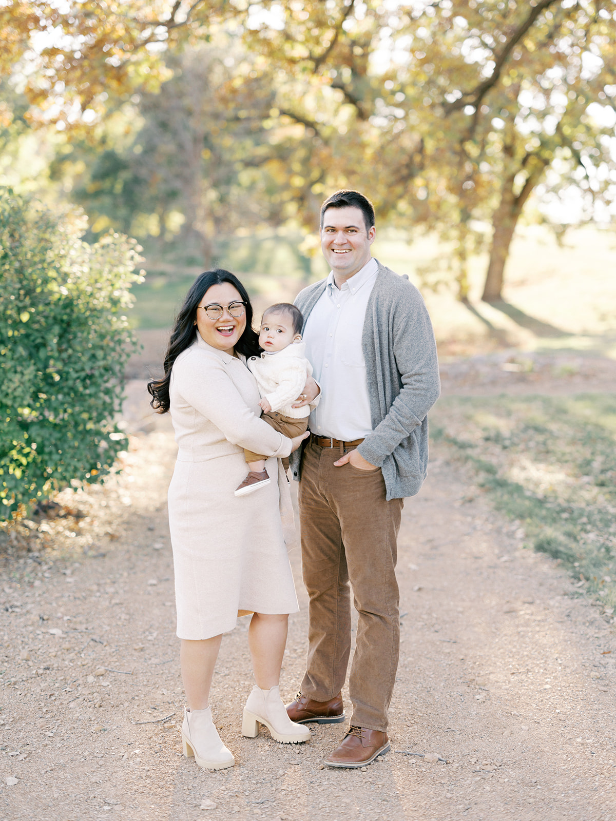 Happy mom and dad stand in a park trail smiling while holding their baby son at one of the photoshoot locations in Minneapolis