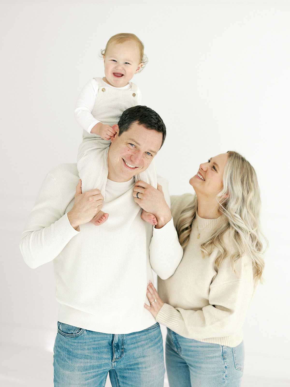 A baby giggles while sitting on dad's shoulders with mom at their side in a studio