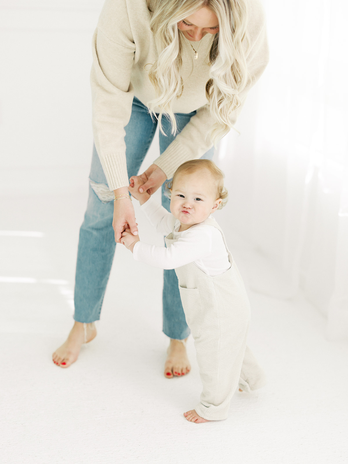 A happy baby walks with mom's help in a white studio in white overalls during some toddler activities in St Paul, MN