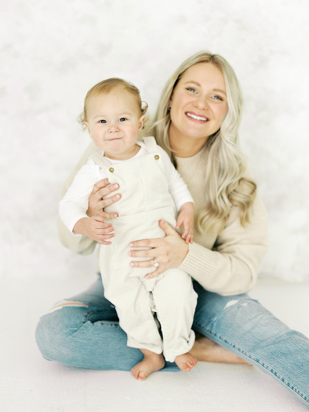 A happy mom sits in a studio in jeans helping her baby in overalls stand with her during some toddler activities in St Paul, MN